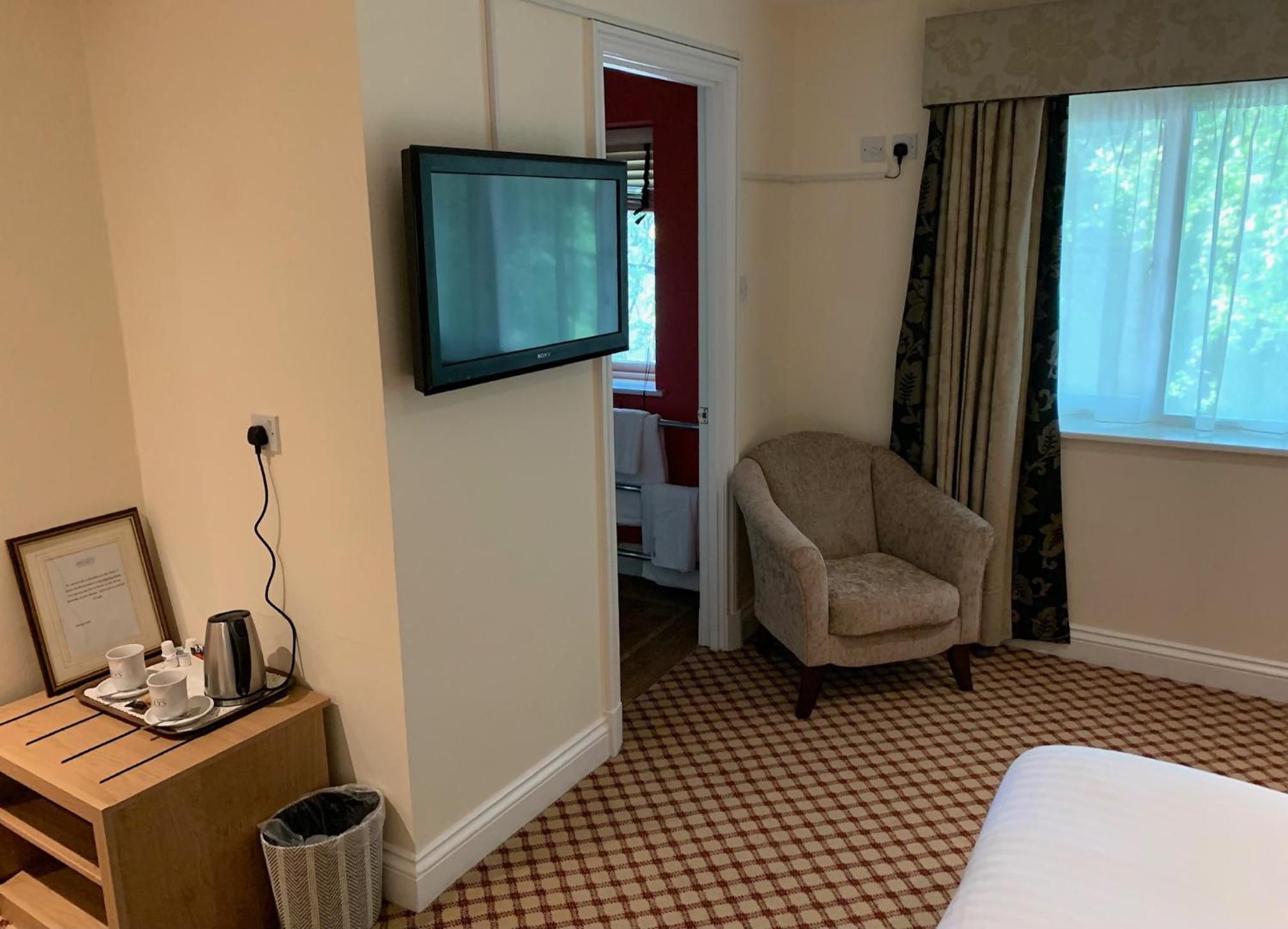 A hotel room with a bed, a television mounted on the wall, a chair, a small table with a tray of cups and a coffee pot, and a checkered floor.