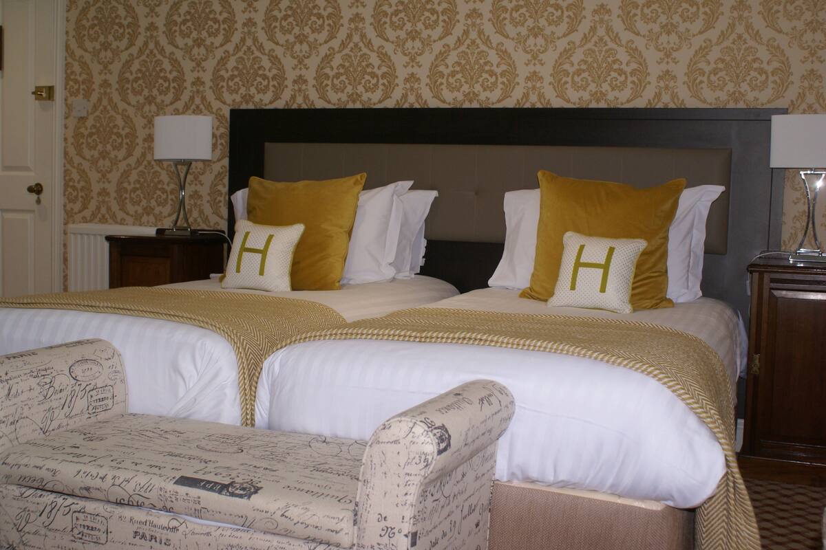 A hotel room with two beds, each with white bedding and yellow pillows, a headboard, a nightstand, and a chair with a vintage design.