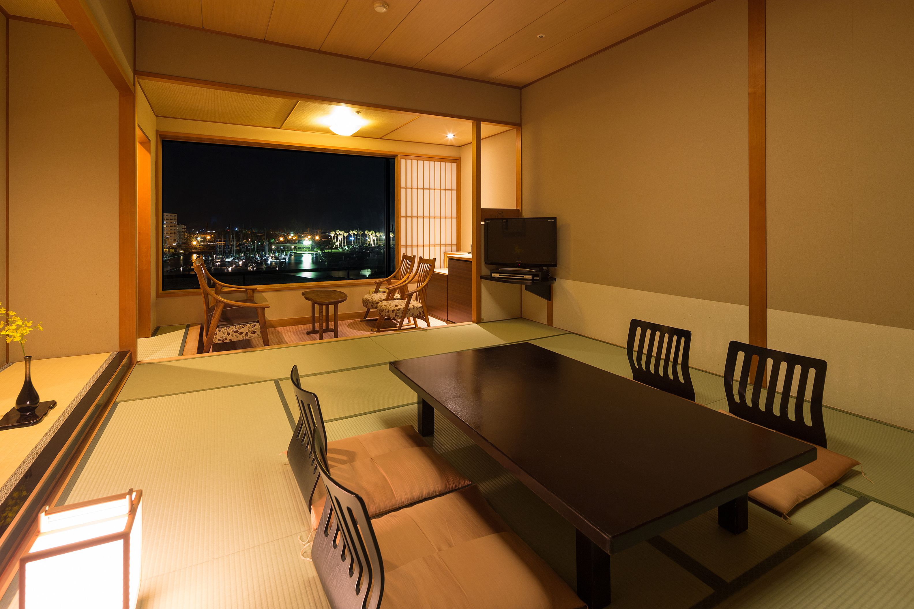 A traditional Japanese room with tatami flooring, a low table, and chairs. A TV is mounted on the wall, and there is a view of a cityscape at night through a large window.