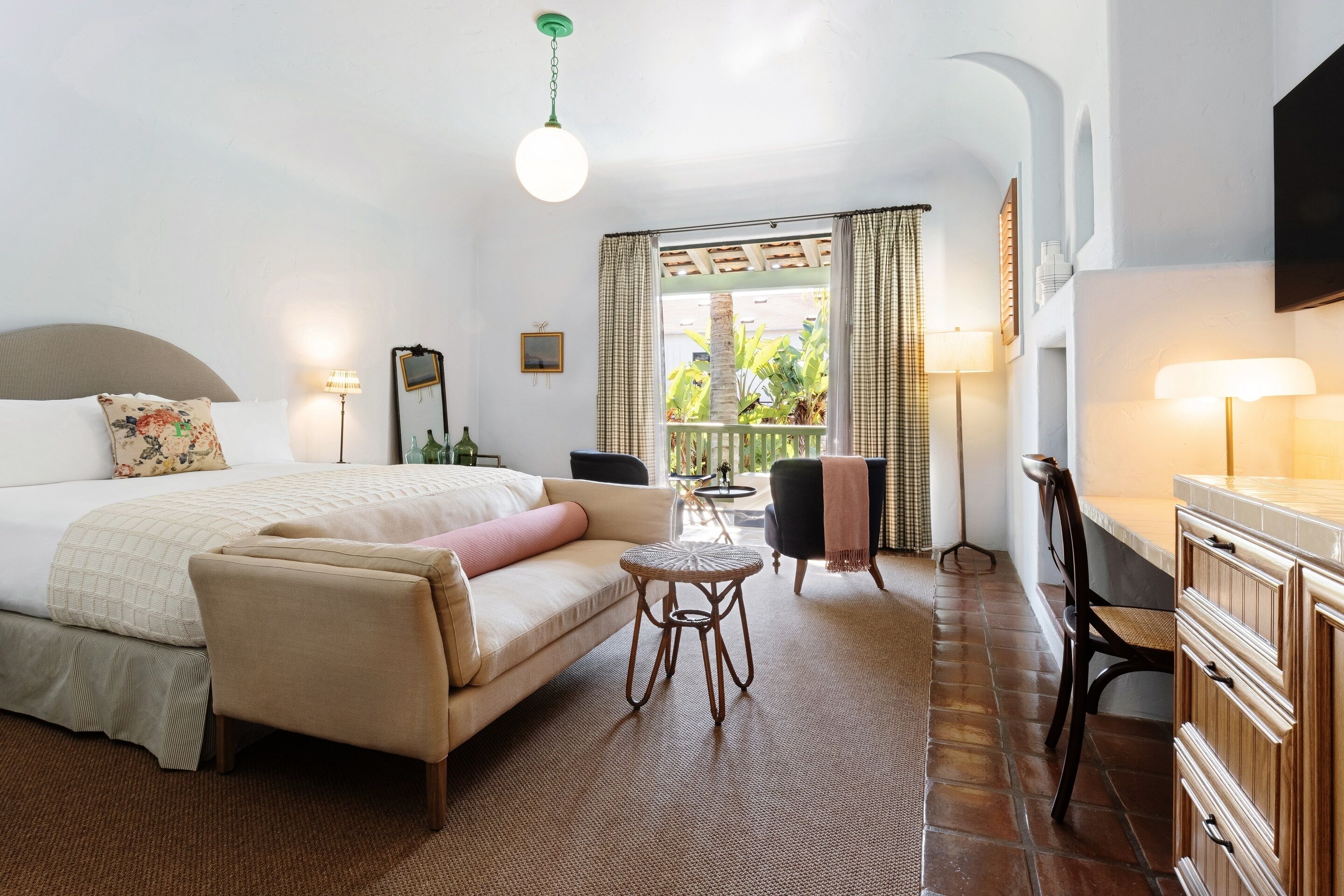 A hotel room with a bed, a sofa, a small round table, a desk with a chair, and a balcony with curtains.