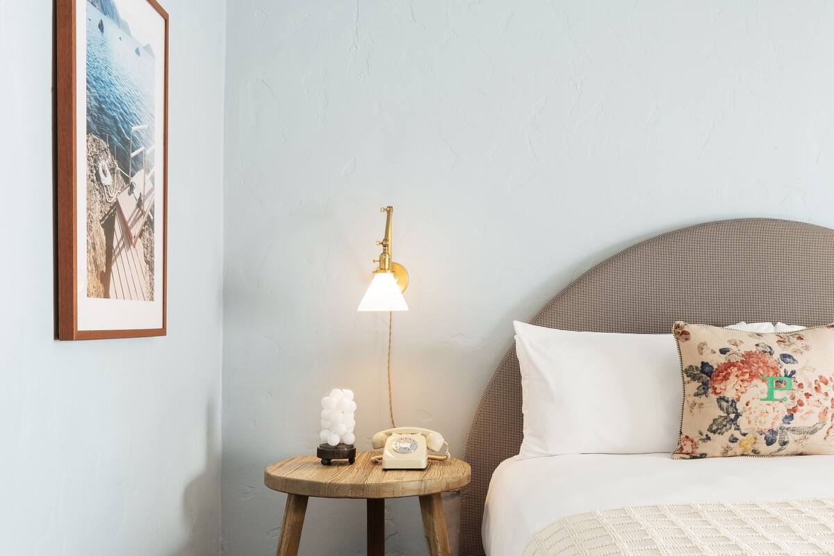 Ein Schlafzimmer mit einem Bett, einem Nachttisch mit Lampe, einem gerahmten Bild an der Wand und einem gemusterten Kissen.