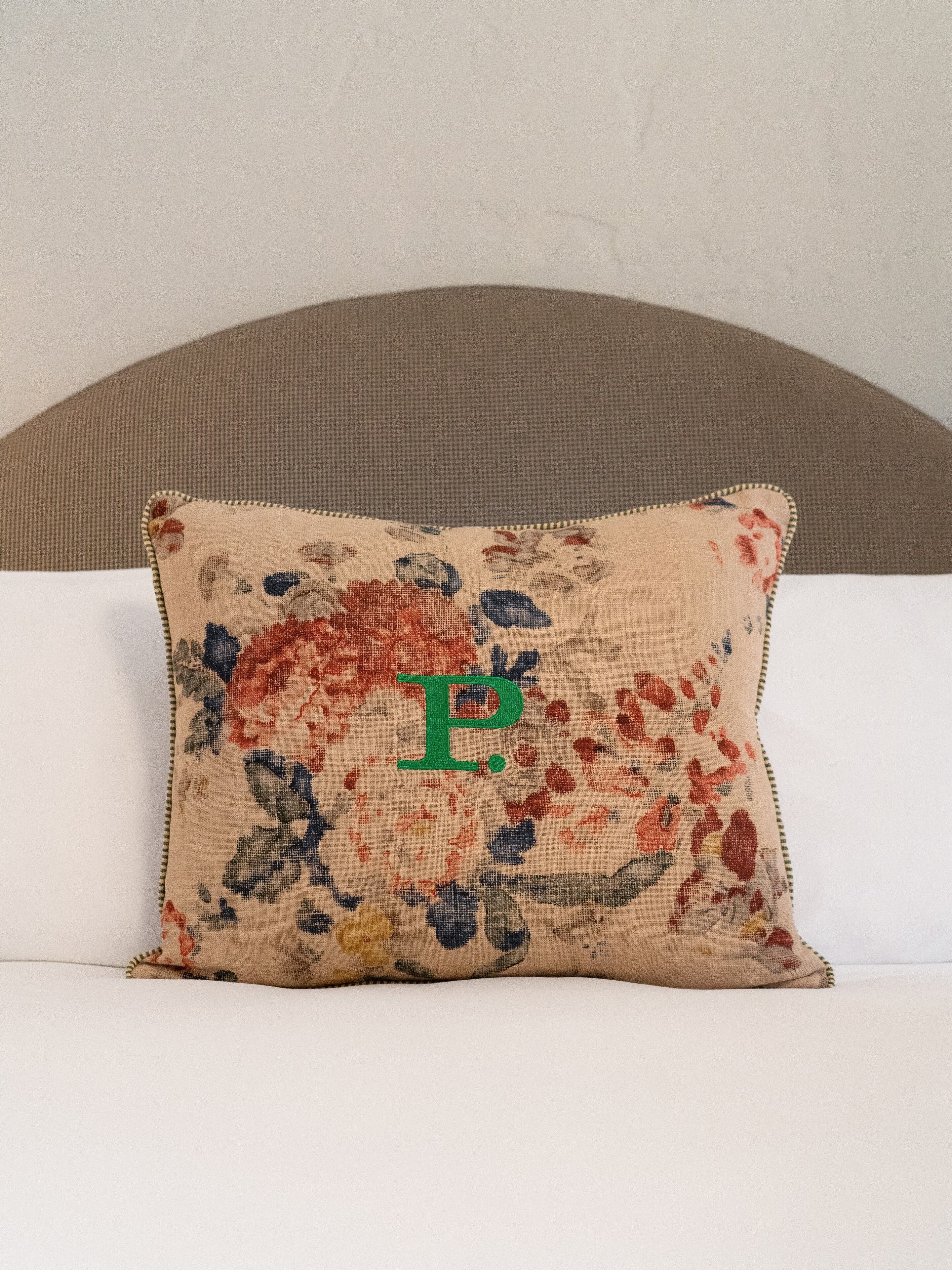 A square pillow with a floral pattern and a large green letter "P" on a bed with a headboard.