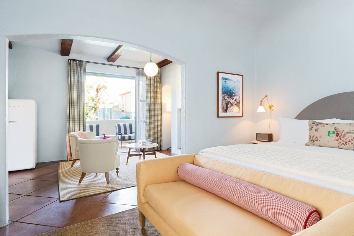 Ein Hotelzimmer mit Bett, Sofa, einem kleinen Tisch und einem Sessel. Es gibt ein Fenster mit Vorhängen, eine Lampe und ein Bild an der Wand.