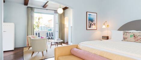 Habitación de hotel con cama, sofá, una mesita y una silla. Tiene una ventana con cortinas, una lámpara y un cuadro en la pared.