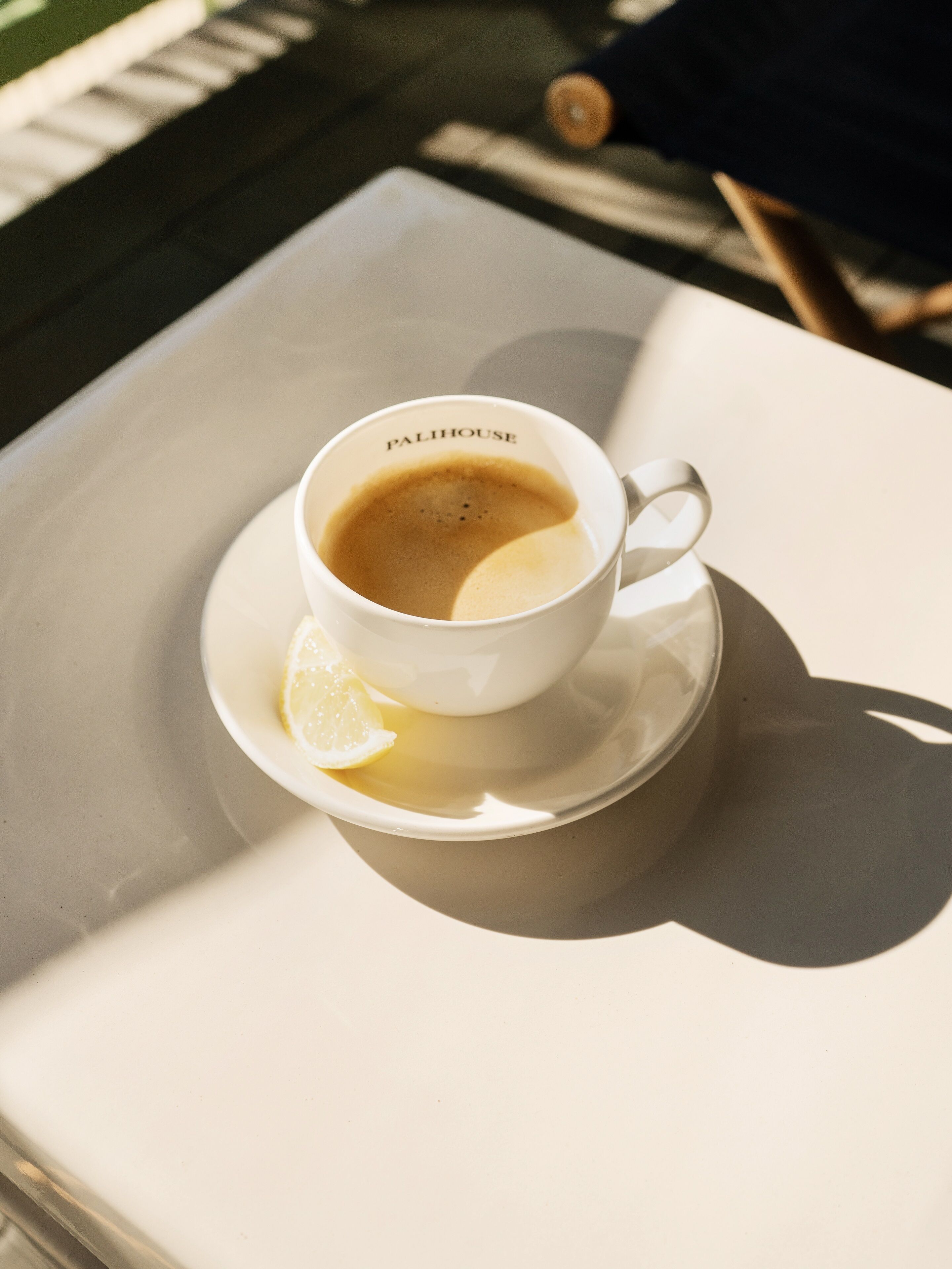 A white cup of coffee with a lemon slice on a white saucer, labeled "PALIHOUSE," on a white table.