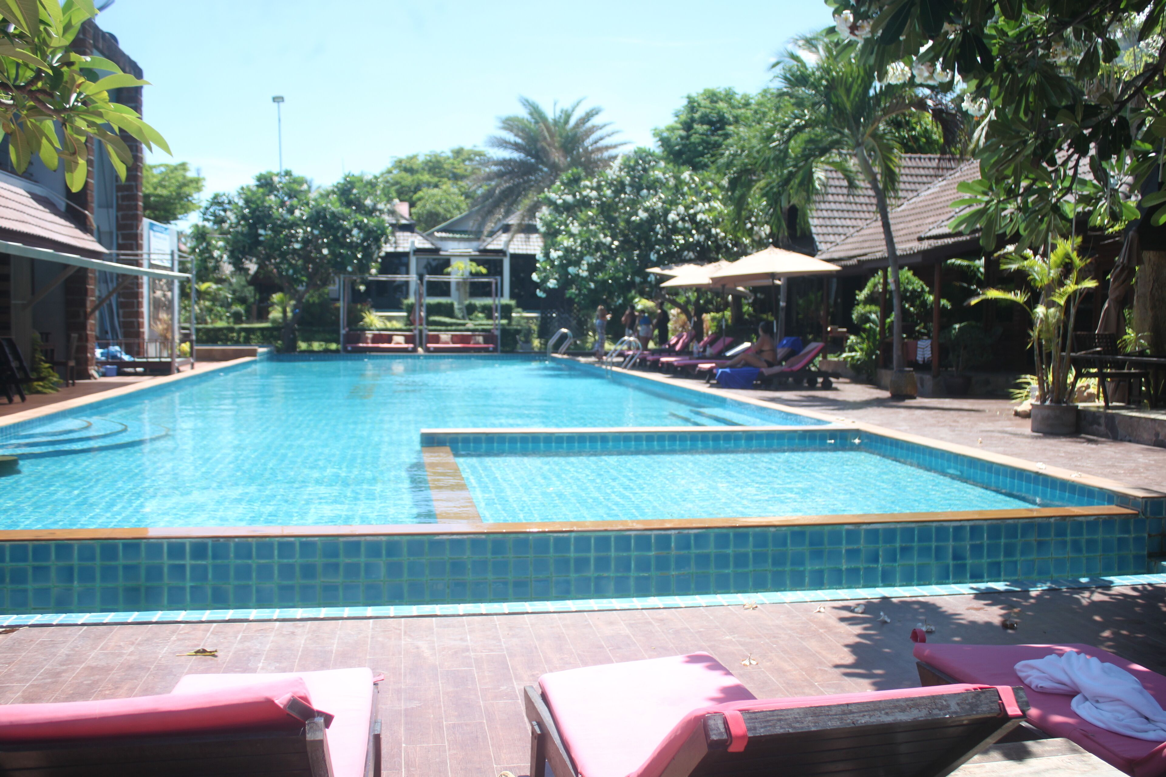 A swimming pool with tiled edges, surrounded by lounge chairs and umbrellas, with a building in the background.