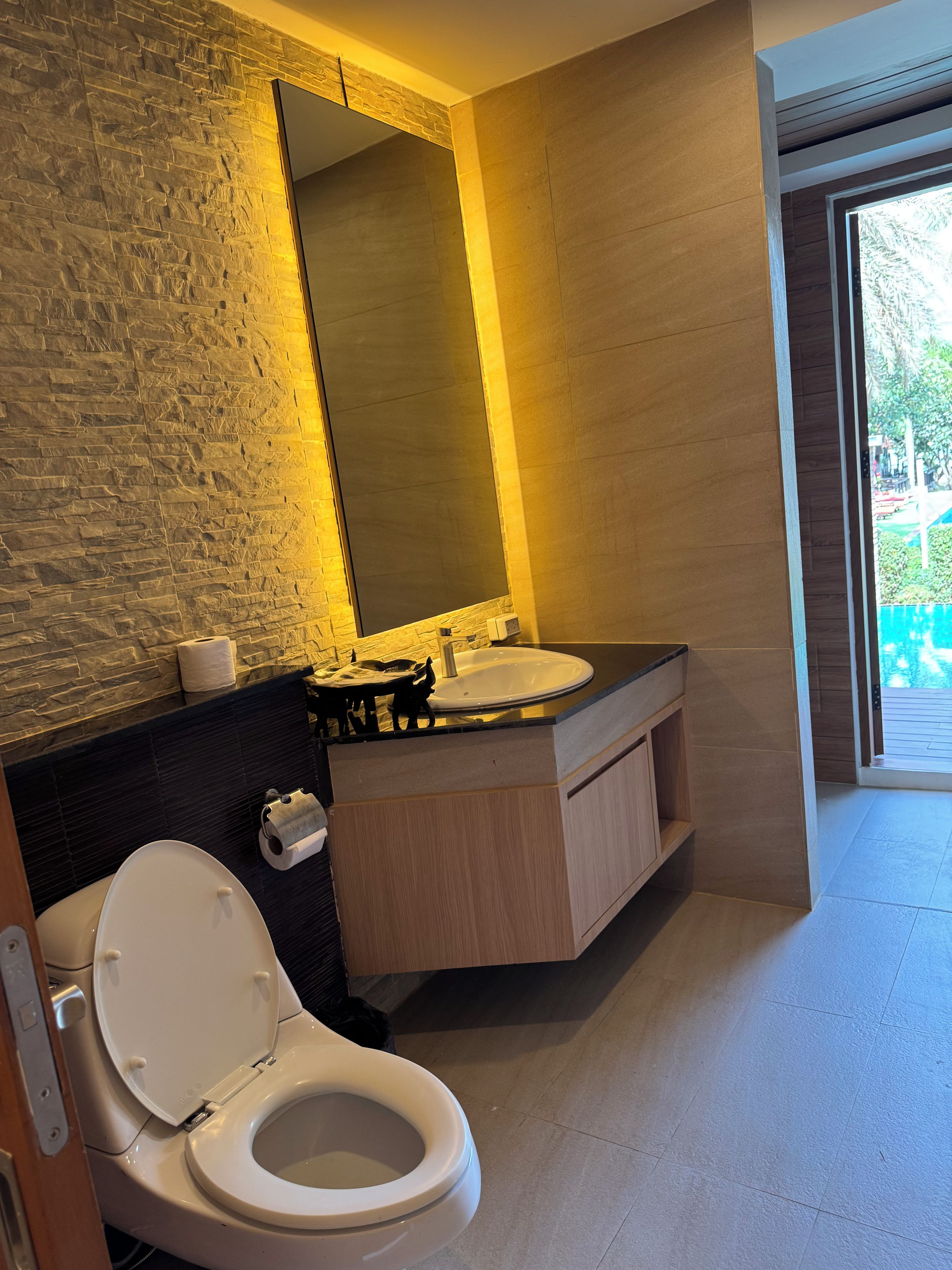 A modern bathroom with a stone-textured wall, a large mirror, a toilet, and a sink with a wooden vanity.