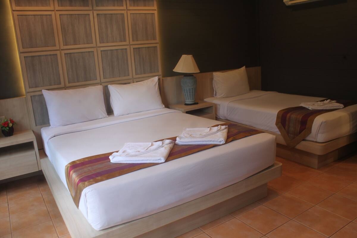 Habitación de hotel con una cama grande, dos camas más pequeñas, una mesita de noche con una lámpara y una mesilla con un jarrón de flores.