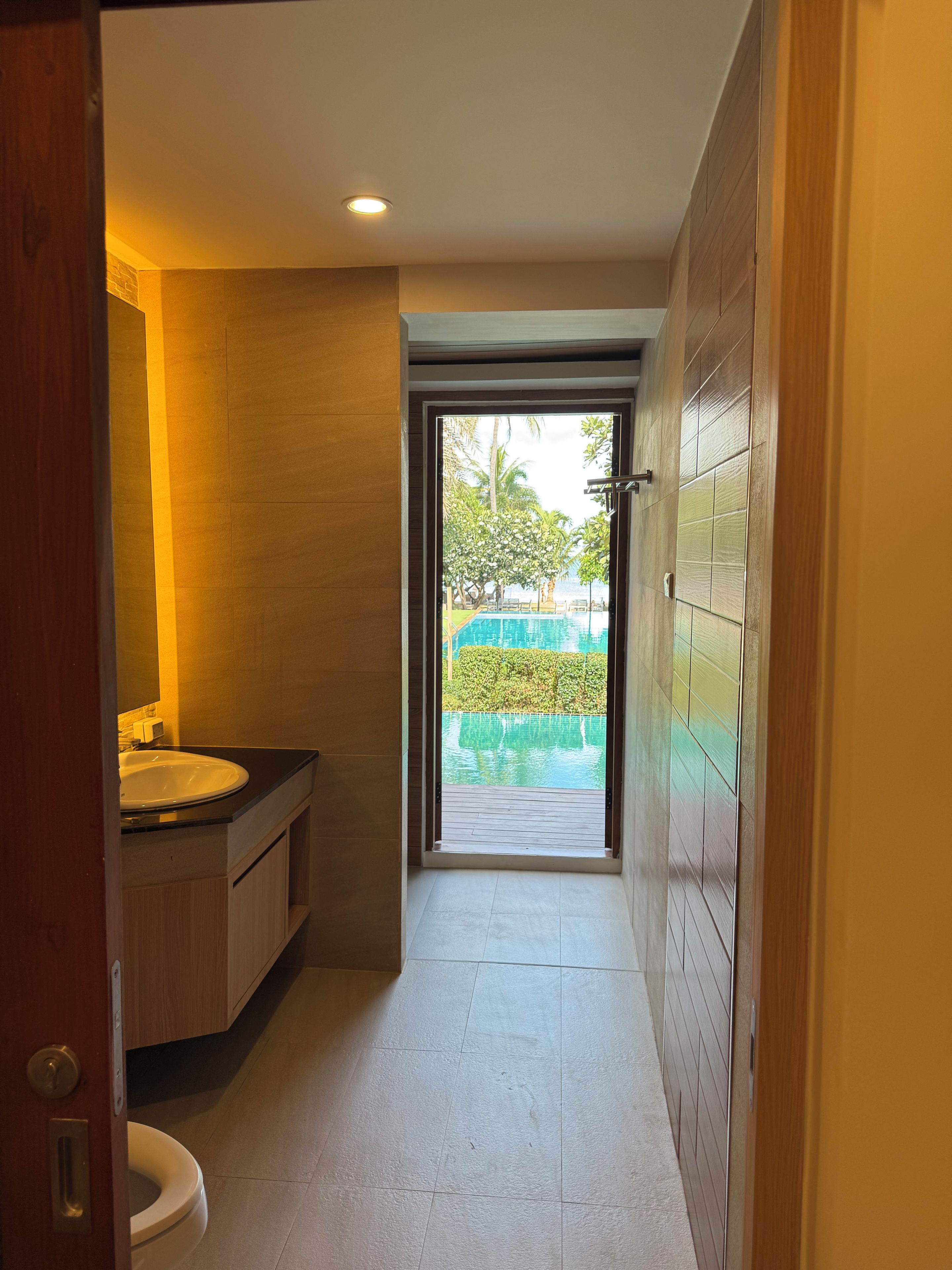 A narrow bathroom with a sink, toilet, and a view of an outdoor pool area.