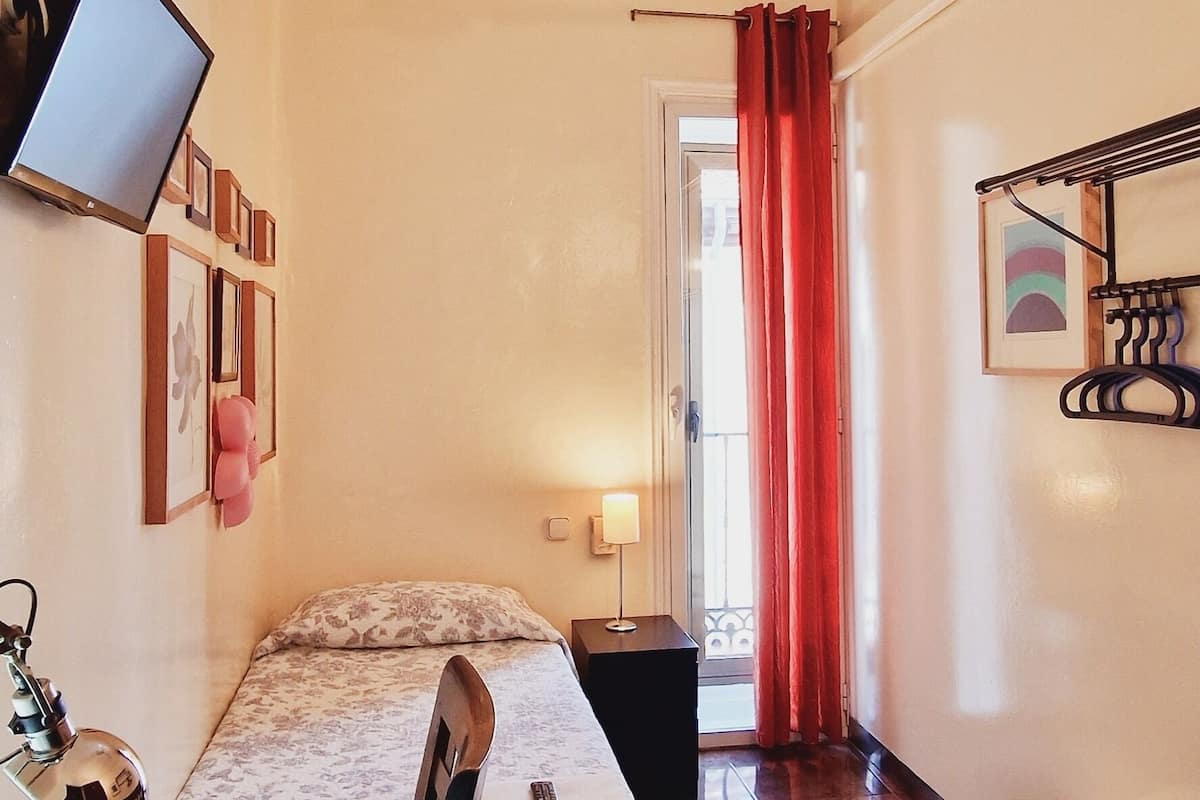 A hotel room with a bed, a desk, a chair, a ceiling fan, a TV, a lamp, and a window with curtains.
