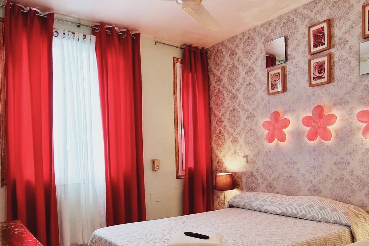 A hotel room with a bed, a desk, a chair, a ceiling fan, a bathroom, and a wall with framed pictures.