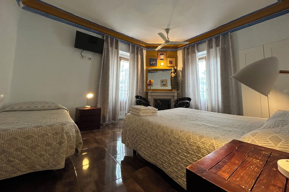 A hotel room with two beds, a ceiling fan, a TV mounted on the wall, a small table with a lamp, and a fireplace.