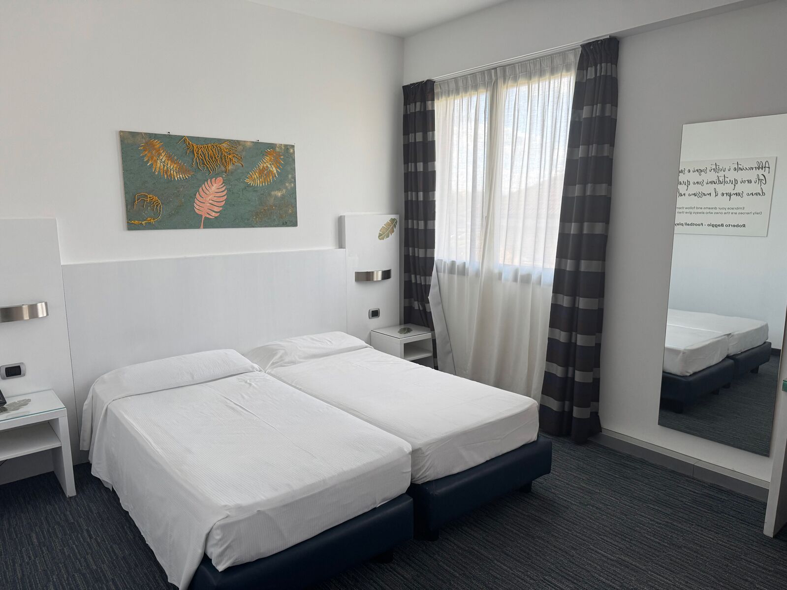 Habitación de hotel con cama doble, mesita de noche, una obra de arte en la pared, ventana con cortinas y un espejo.
