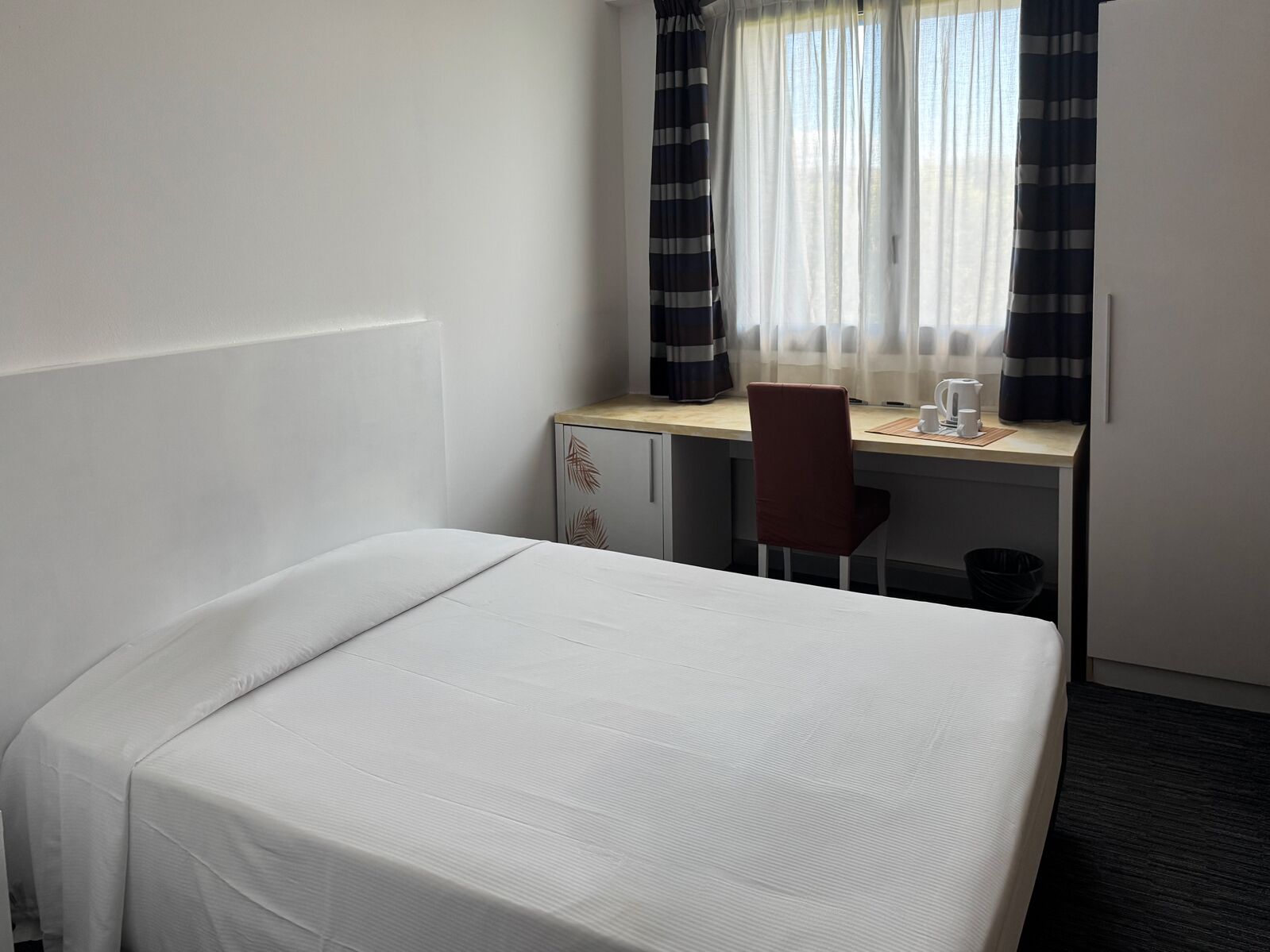 Quarto de hotel com uma cama de solteiro, uma escrivaninha, uma cadeira e uma janela com cortinas.