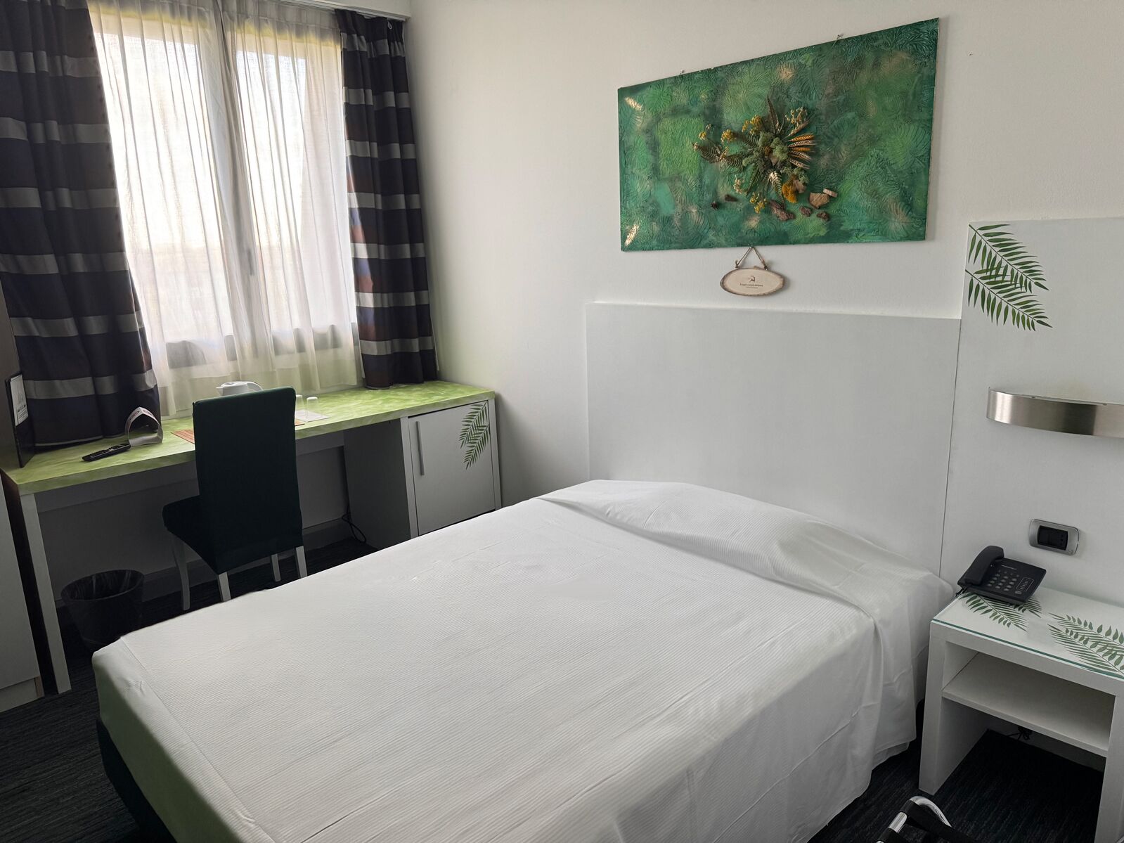 Habitación de hotel con cama, escritorio, silla, una mesita con teléfono y un cuadro en la pared.