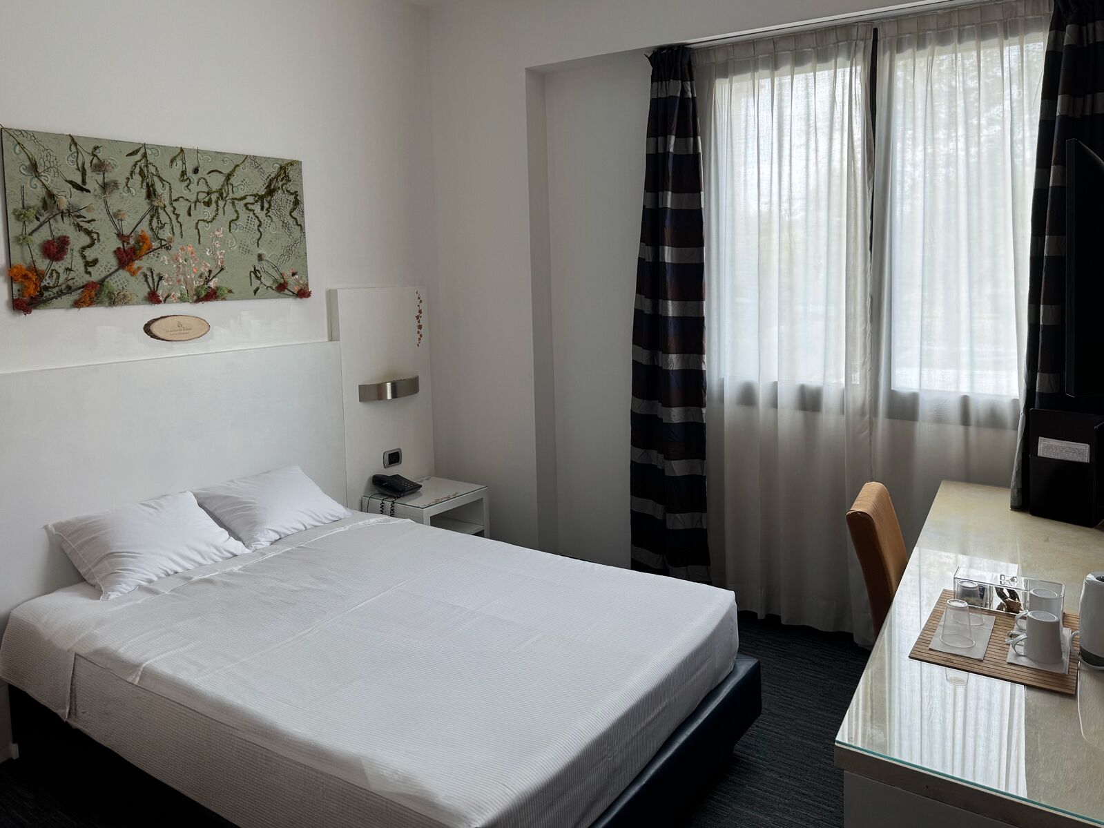 Ein Hotelzimmer mit Bett, Nachttisch, Schreibtisch mit Stuhl, Spiegel und einem fensterbepflanzten Fenster.