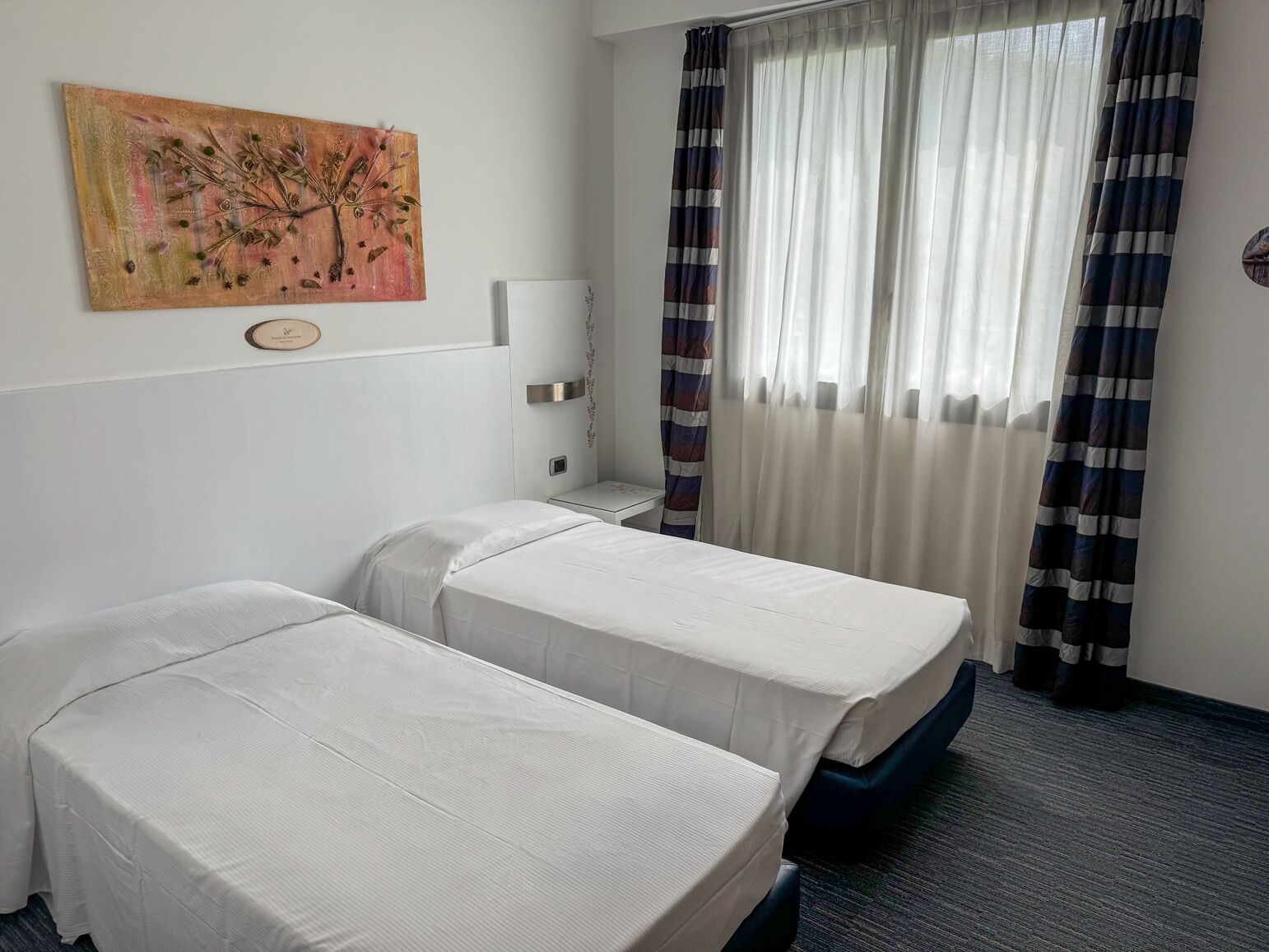 Habitación de hotel con dos camas individuales, un cuadro en la pared, una mesita de noche y un ventanal con cortinas.