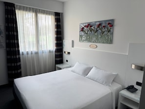 A hotel room with a bed, a bedside table with a phone, a nightstand, a painting on the wall, and a window with curtains. - B&B HOTEL Milano Cologno Sporting (Cologno Monzese)