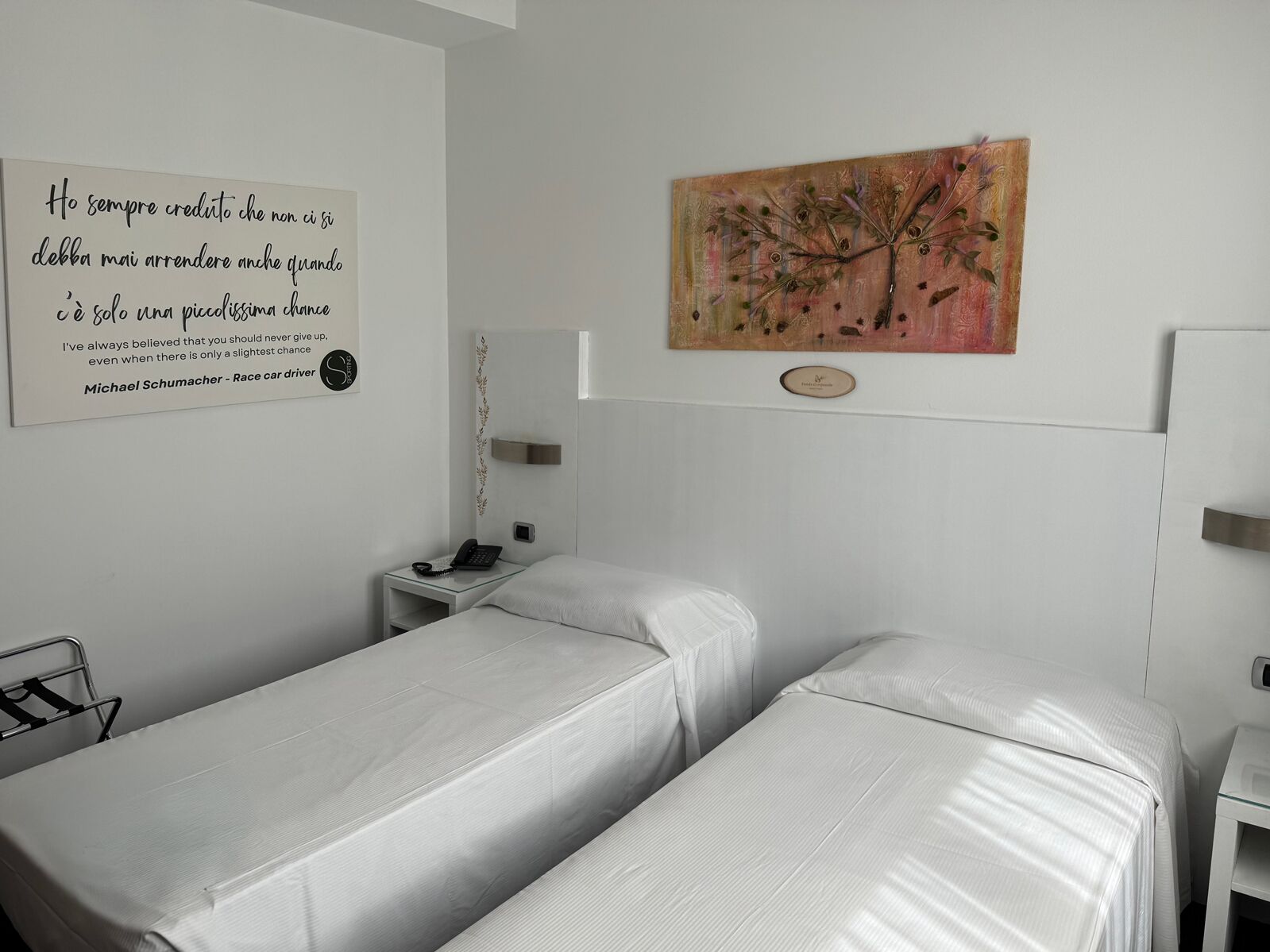 Una habitación de hotel con dos camas, una mesita de noche con un teléfono, una mesita de noche pequeña con una lámpara, y una pared con una cita y un cuadro.