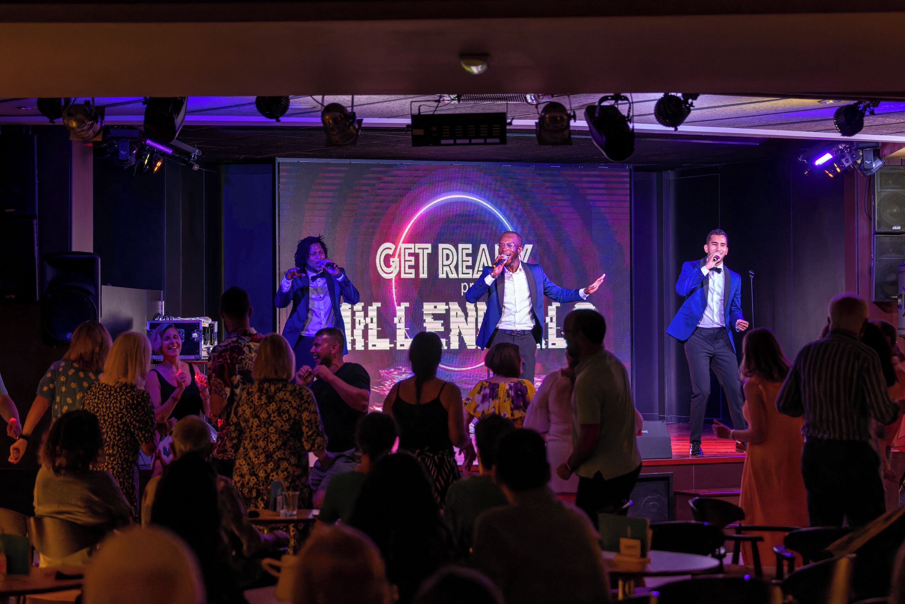 Un escenario con una pantalla grande que muestra los textos 'GET REAL' y 'KILL IT', rodeado por un público en un local con poca luz.