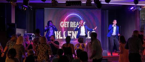 A stage with a large screen displaying "GET REAL" and "KILL IT" text, surrounded by an audience in a dimly lit venue.