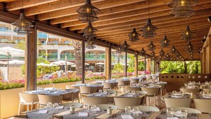 Un área de comedor al aire libre con varias mesas dispuestas para comensales, con techo de madera y amplias ventanas con vistas a un jardín ajardinado.