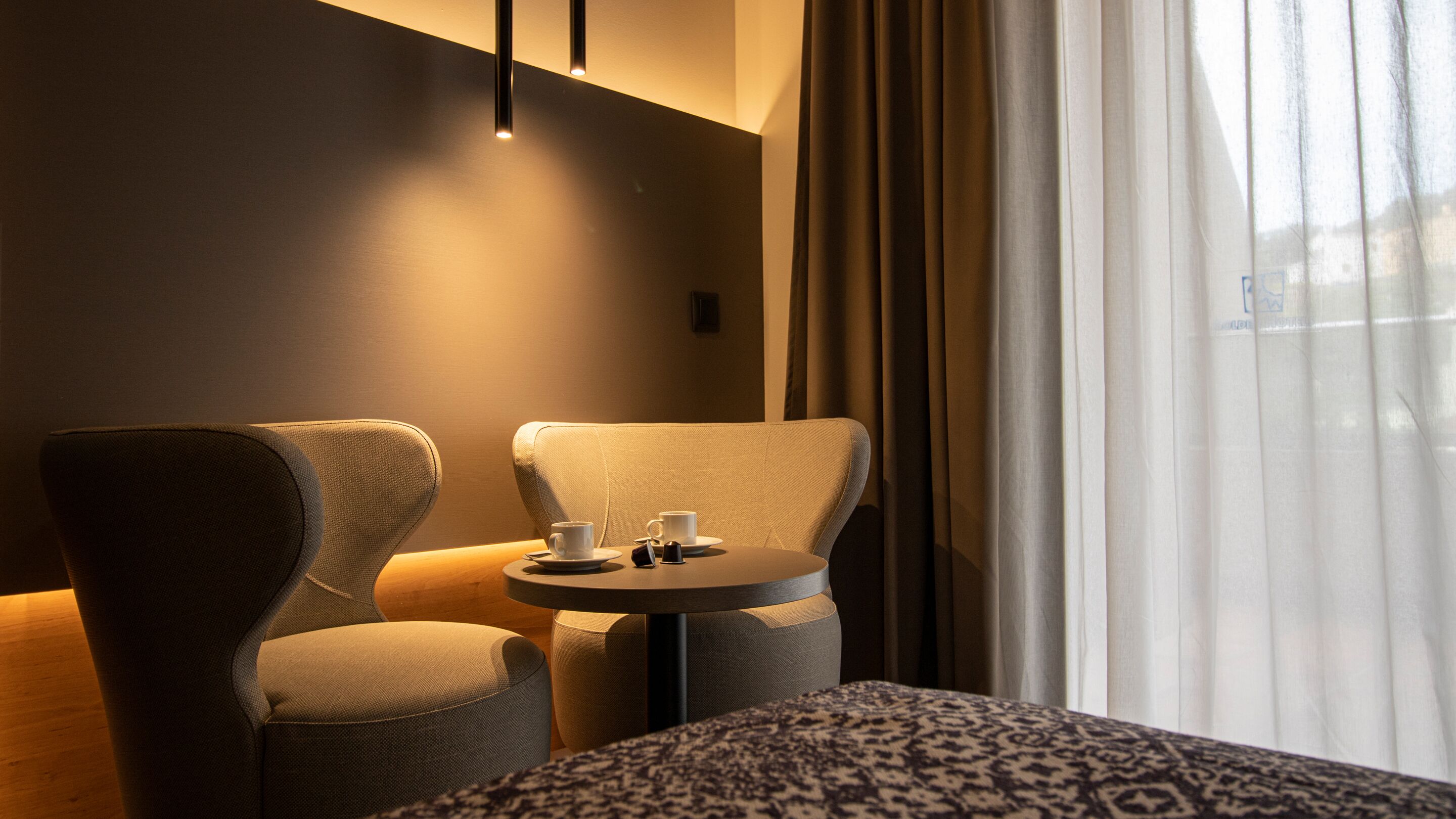 Une chambre d’hôtel avec deux fauteuils, une petite table ronde sur laquelle se trouvent une tasse et une soucoupe, et un lit recouvert d’une couverture à motifs.