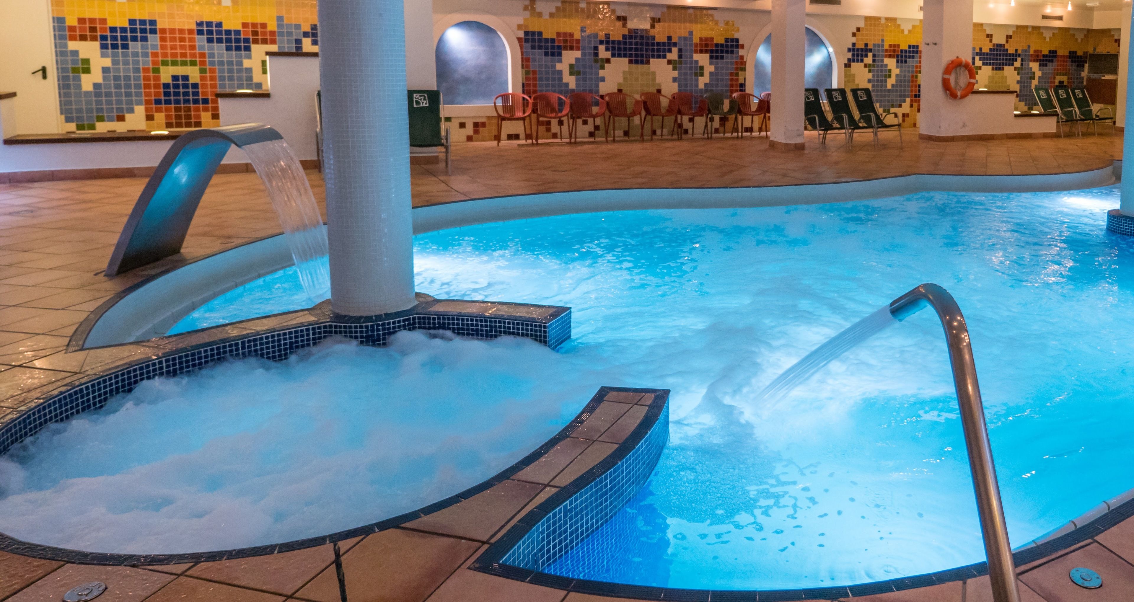 An indoor swimming pool with a hot tub area, lounge chairs, and a mosaic wall.