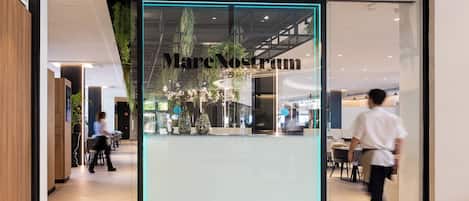 A modern restaurant interior with a glass entrance, indoor plants, and a visible seating area.