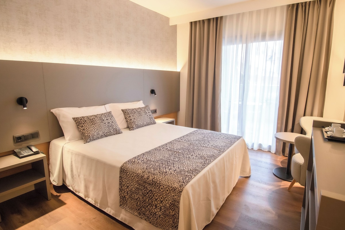 Une chambre d’hôtel avec un grand lit, des tables de chevet, un bureau avec une chaise, une machine à café et une fenêtre avec des rideaux.