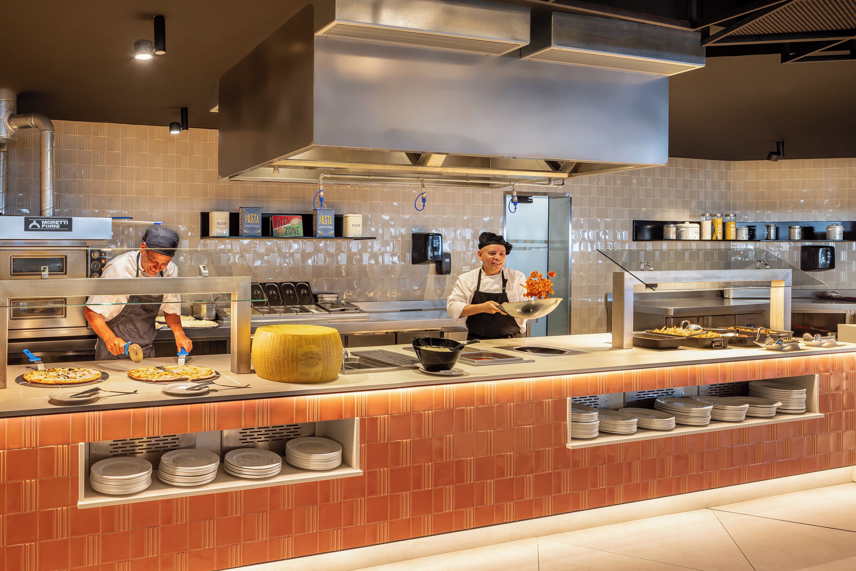 A modern kitchen with two chefs preparing pizzas, a large yellow cheese wheel, and various kitchen utensils and ingredients on the counter.