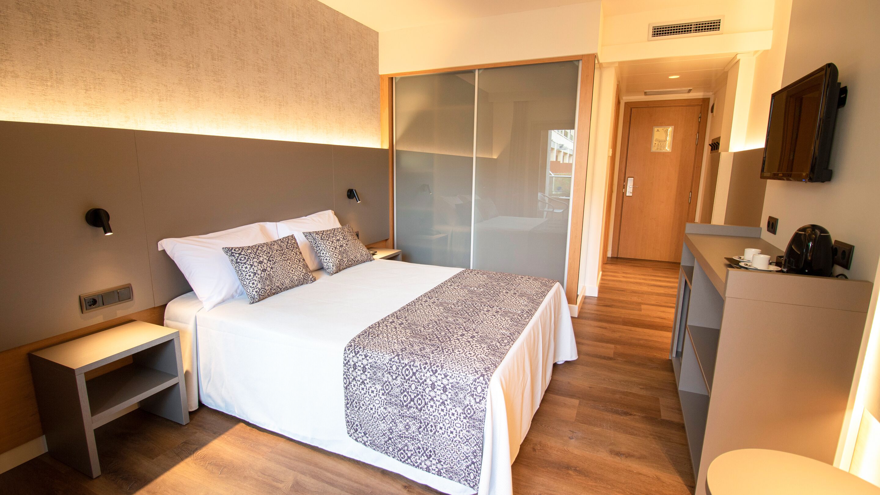 Habitació d'hotel moderna amb un llit gran, taules de nit, un armari, un escriptori amb una màquina de cafè i un mirall.