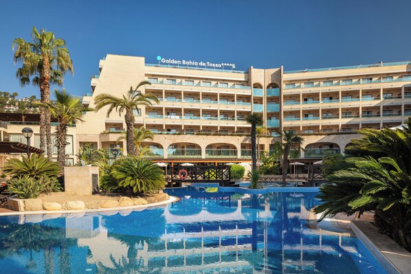 Een hotel met een groot zwembad, palmbomen en een helderblauwe lucht.