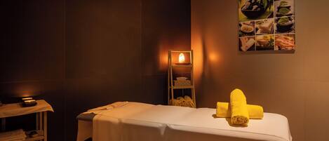 A spa room with a massage table, towels, a shelf with towels, and a wall art piece.