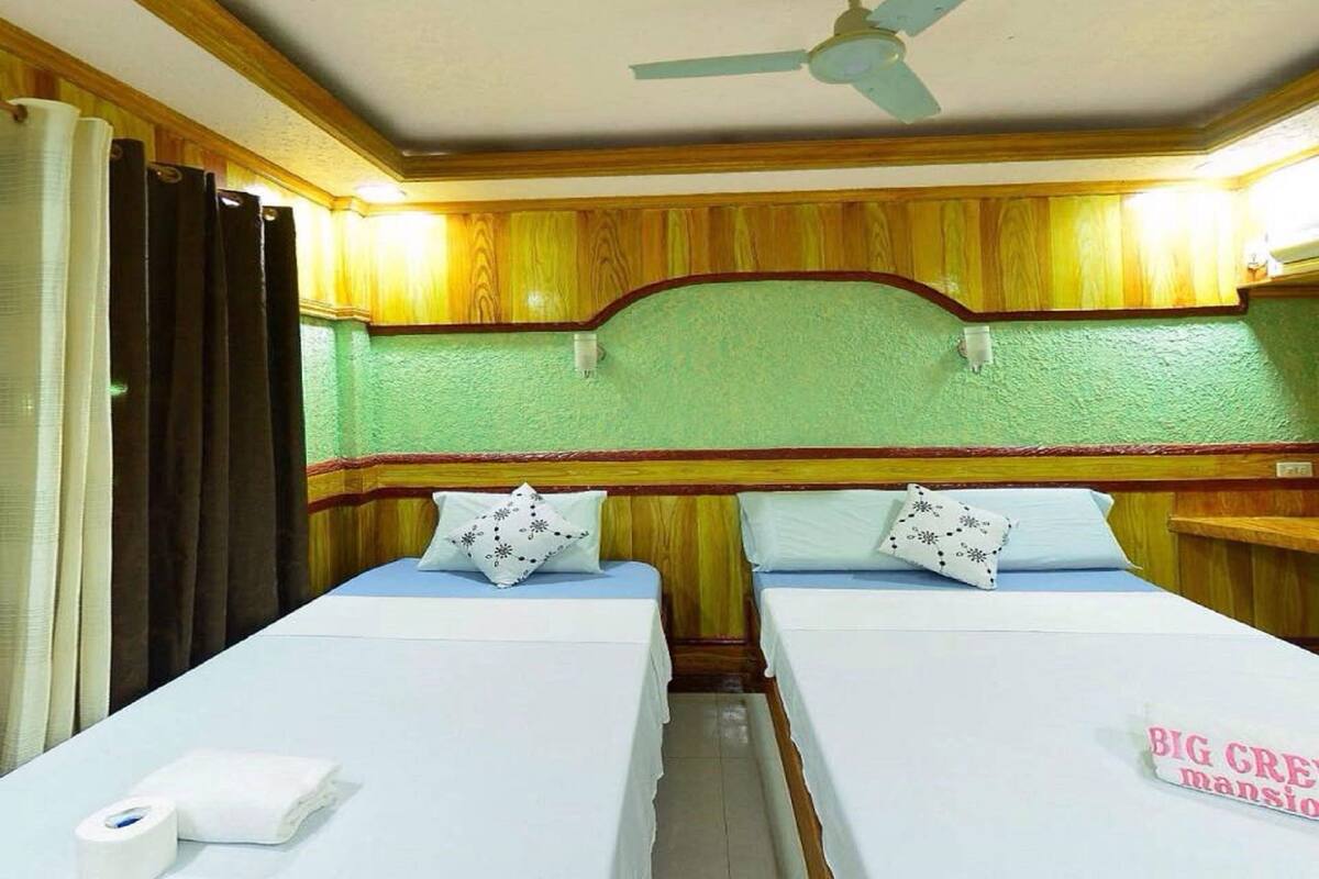 Une chambre avec deux lits, une tête de lit en bois, un ventilateur de plafond et un panneau accroché au mur.