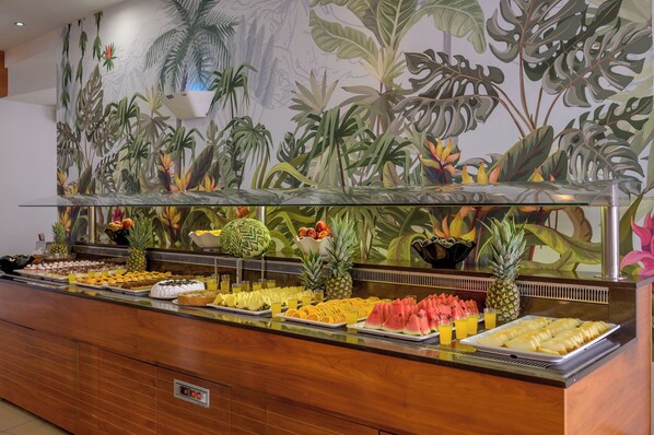 A buffet with tropical-themed wall art, a variety of fruits, and a digital clock.