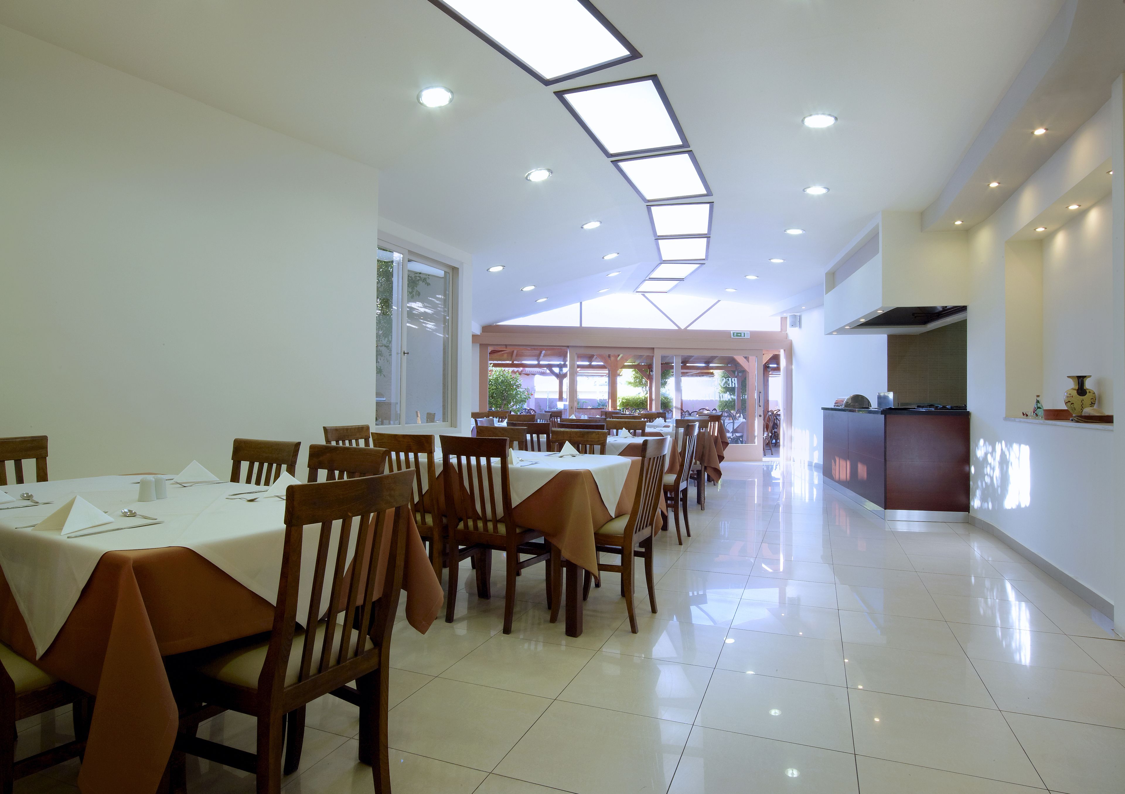 A spacious dining area with multiple tables and chairs, a kitchen area, and large windows allowing natural light.
