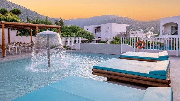 A pool with a water feature, lounge chairs, and a view of buildings and mountains. - Eurohotel Katrin Hotel & Bungalows (Hersonissos)