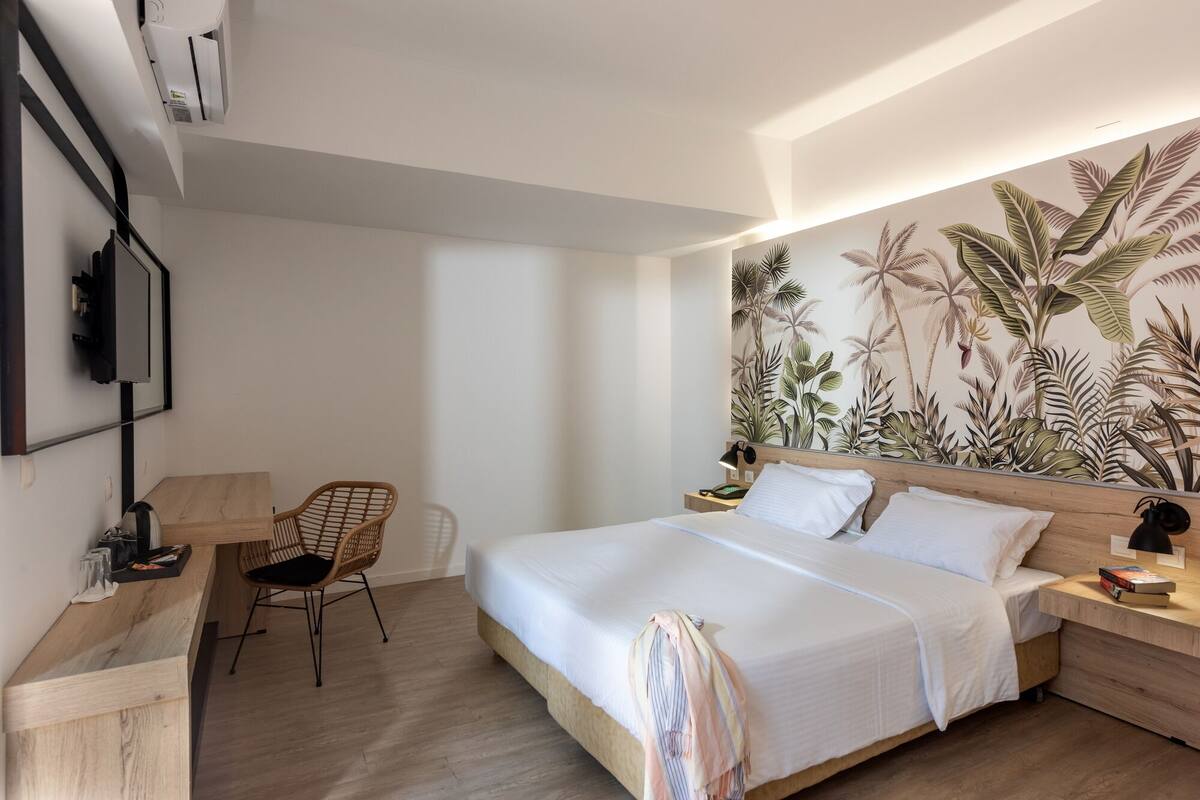 A modern hotel room with a large bed, a desk with a chair, a TV, and a wall with a tropical plant mural.