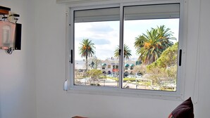 Habitación de hotel con dos camas, una ventana que ofrece vista a palmeras y un estacionamiento, y una pared blanca con toma de corriente.