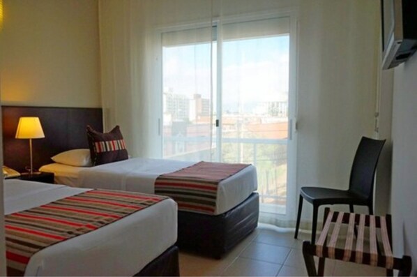 A hotel room with two beds, a chair, a TV, and a window with a city view. - De la Plaza Hotel (Maldonado)