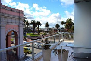A balcony with a view of a street, palm trees, and a building with a pink facade.