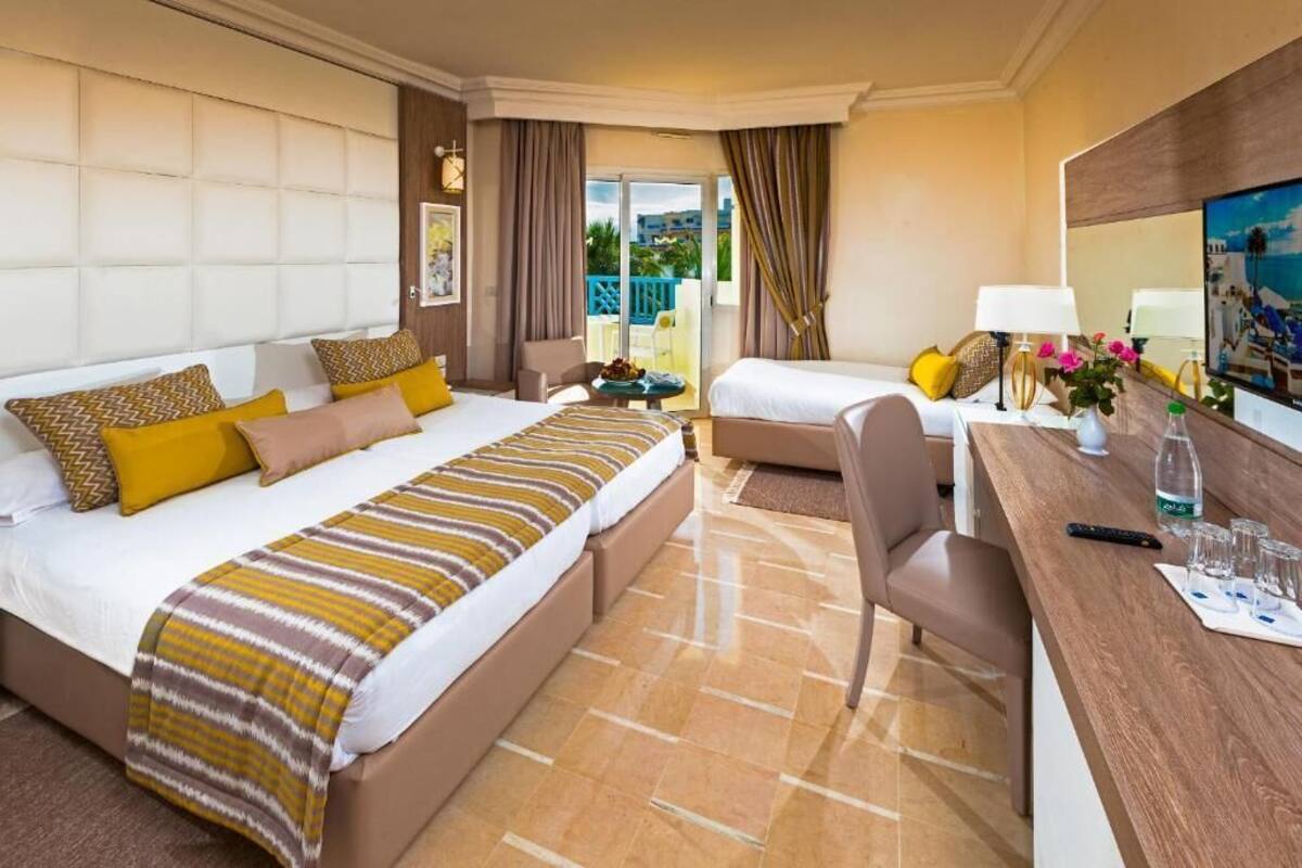 Une chambre d’hôtel avec un grand lit, un lit plus petit, un bureau avec une chaise et un balcon avec des rideaux.