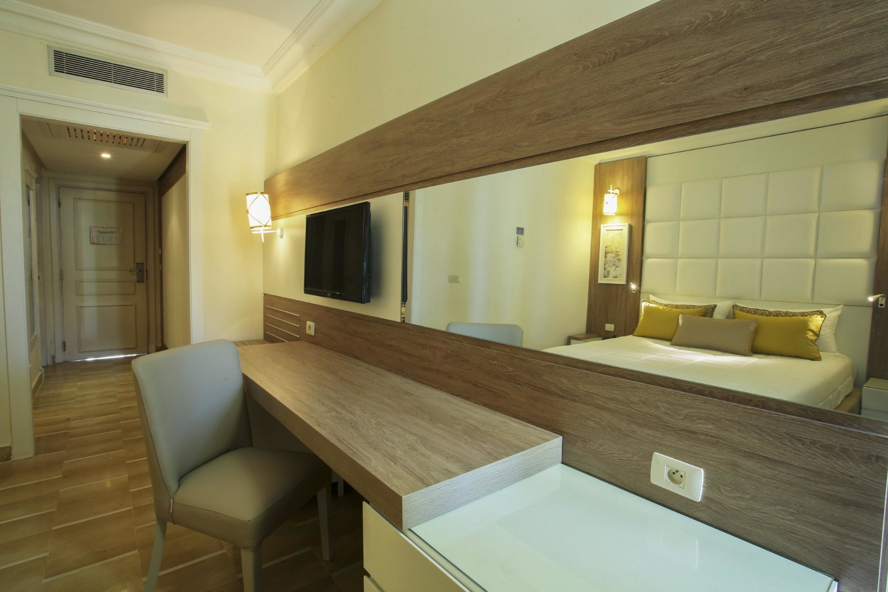 A hotel room with a wooden desk, a chair, a large mirror, a bed with yellow pillows, and a television mounted on the wall.