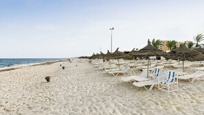 Ein Strand mit weißen Liegestühlen und Reetdachschirmen, einem klaren Himmel und ruhigem Meer.