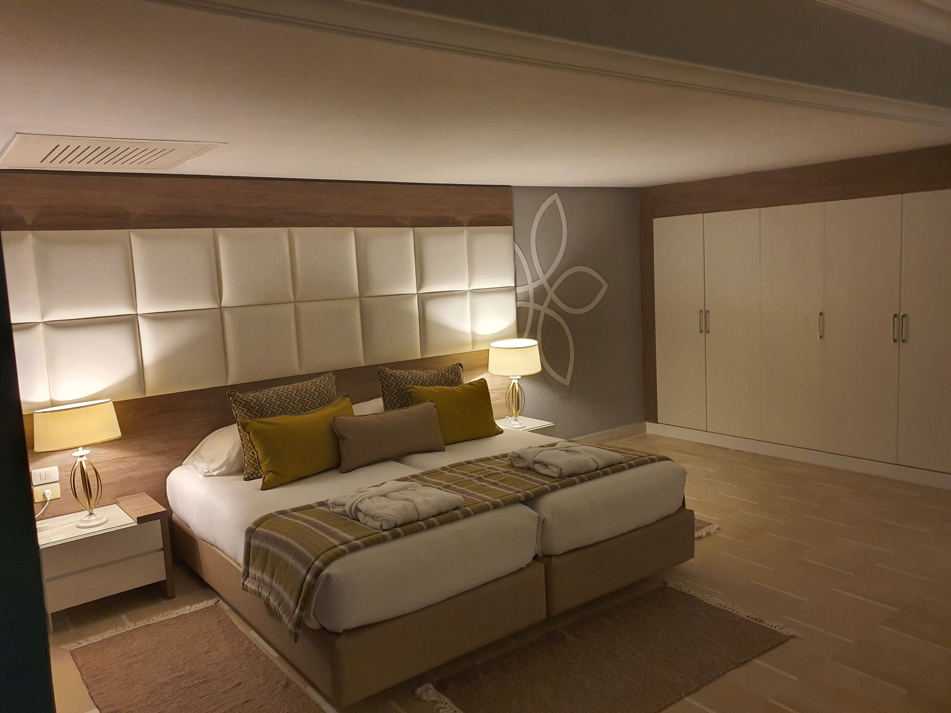 Un dormitorio moderno con cabecera capitonada, una cama grande con muchas almohadas, una mesita de noche con lámpara y un armario.