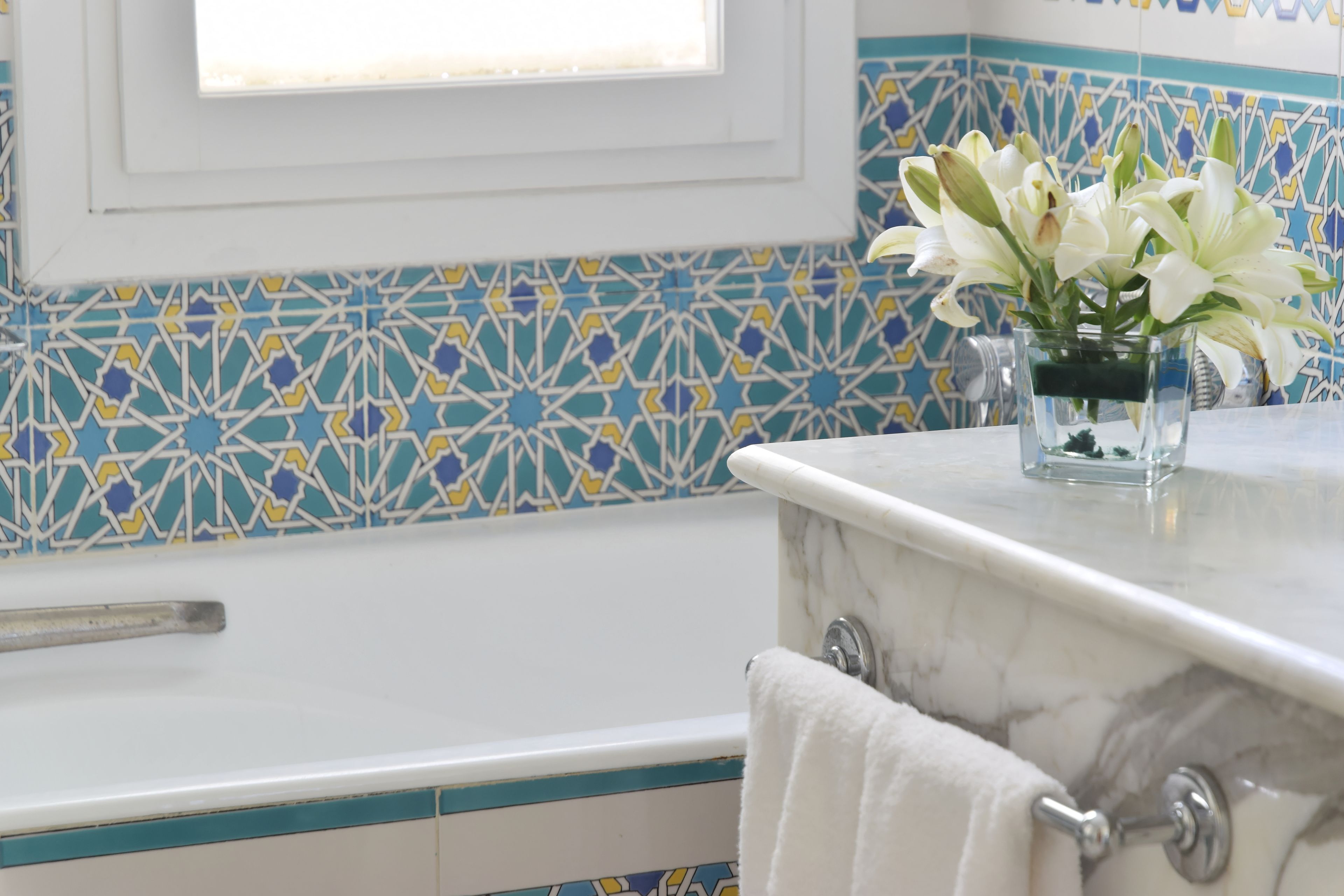 Un baño con una bañera blanca, un florero de vidrio con flores blancas y un salpicadero de azulejos estampados.