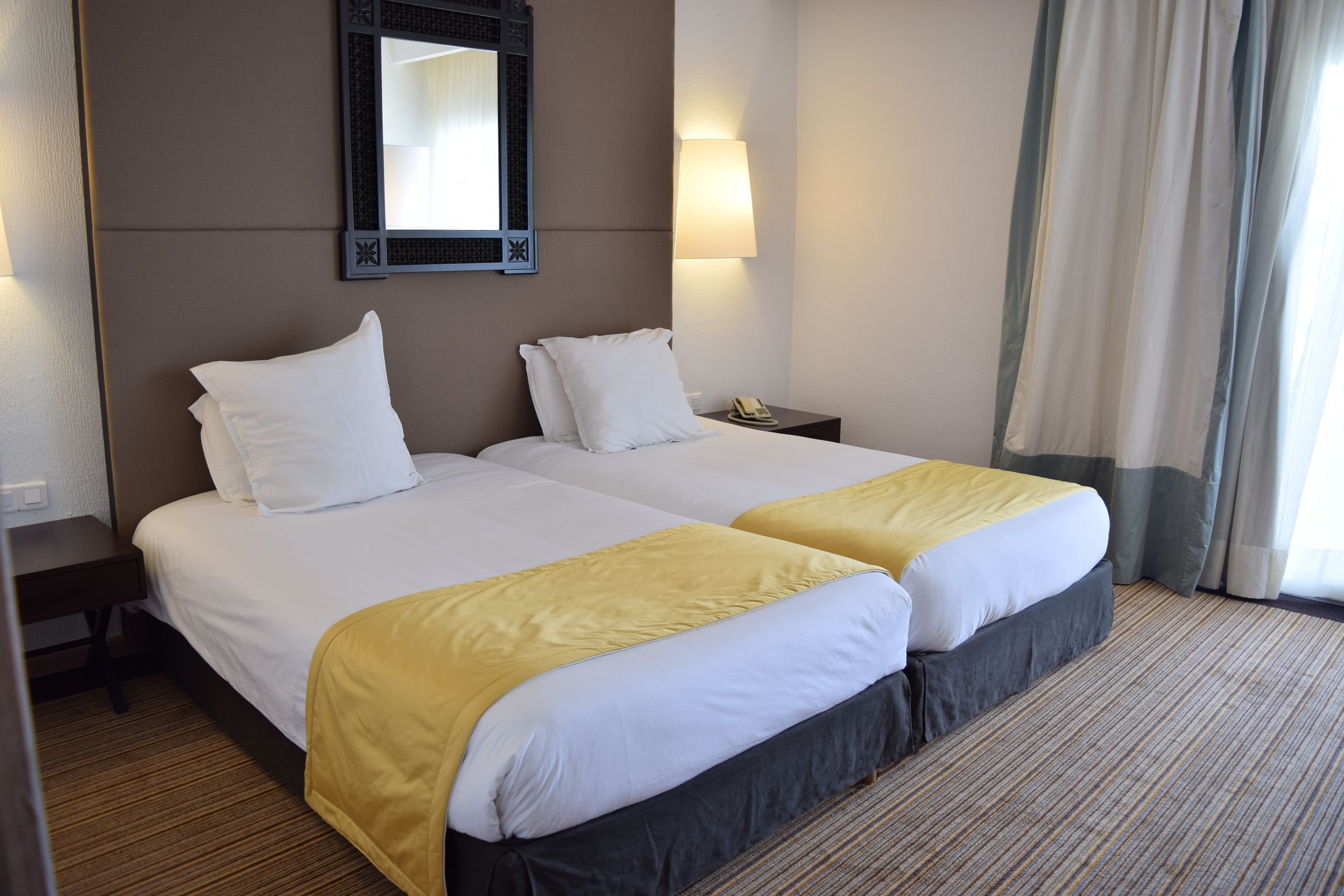 A hotel room with two beds, a nightstand, a mirror, a lamp, and a window with curtains.