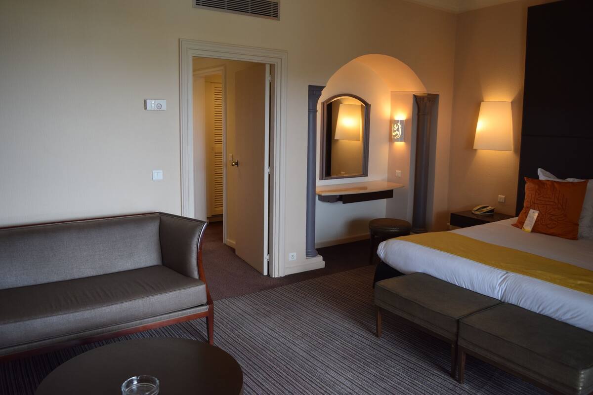 A hotel room with a sofa, a bed with a yellow bedspread, a bench, a nightstand with a lamp, and a mirror above the bench.