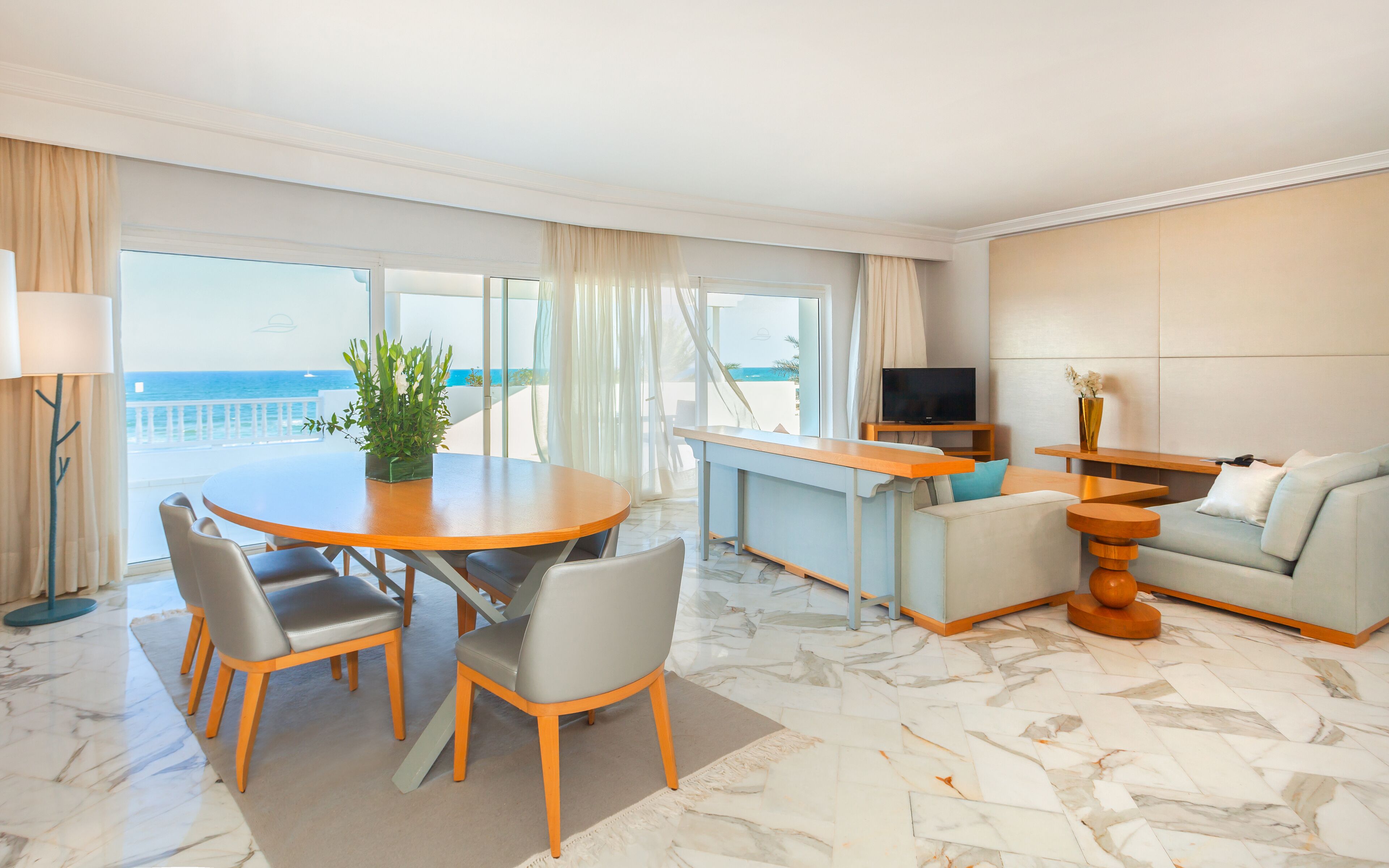 A hotel room with a dining area, a sofa, a TV, and a view of the sea.